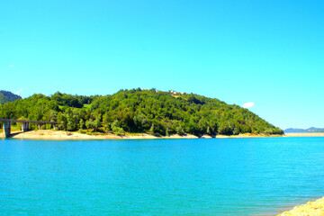View on the shores of Gerosa Lake with its wooly silky blue waters delimited by a dam in the distance, a hill full of green vegetation and a bridge connecting through the wilderness on a summer day