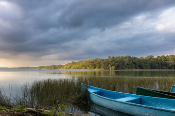 Blue canoe on shore with marshes and fresh water lagoon surrounded by tropical green jungle with light reflection and cloudy sky at sunset