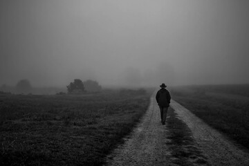 A moody vibe as a man saunters down a dirt road on a calm foggy morning. Man walking away.