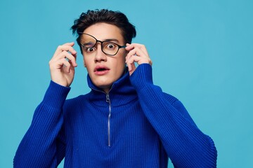 a handsome, attractive man stands on a light blue background in a blue zip-up sweater and holding black-rimmed glasses on his face with his hand, looks in surprise at the camera with his mouth open