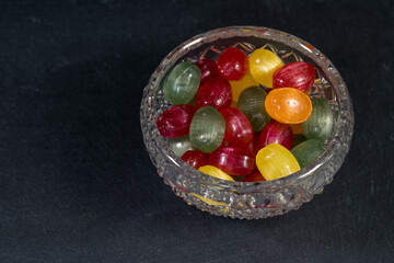 hard candies fruit flavor in various colors (red, green, yellow, orange), in a glass, crystal bowl, candy, sweets, high sugar content, unhealthy snacks, close-up shot, blurred black background