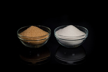 Two types of sugar, white beet sugar and brown cane sugar, in glass bowls, standing on a black reflective background. Comparison of types of sugar and their properties.