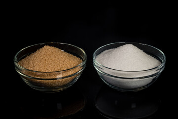 Two types of sugar, white beet sugar and brown cane sugar, in glass bowls, standing on a black reflective background. Comparison of types of sugar and their properties.