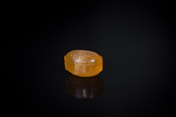 single hard candie fruit flavor orange color, on a black glossy background, candy, sweets, high sugar content, unhealthy snack, close-up shot, blurred background