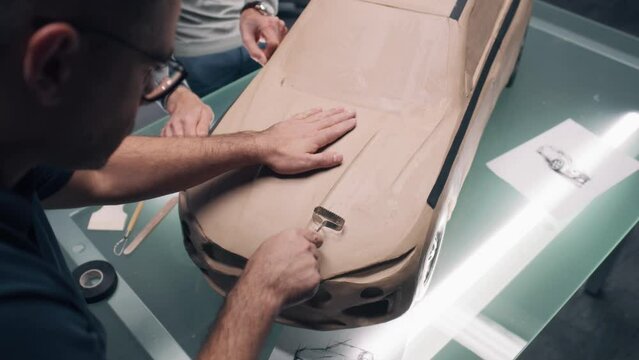 Two male automotive designers sculpting futuristic plasticine clay model of car with rake. Future design of new generation electric car. Working in modern car design studio.