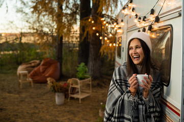 Caucasian woman in a knitted hat wrapped in a plaid and drinks a warming drink outdoors. Travel in a motor home in the fall.