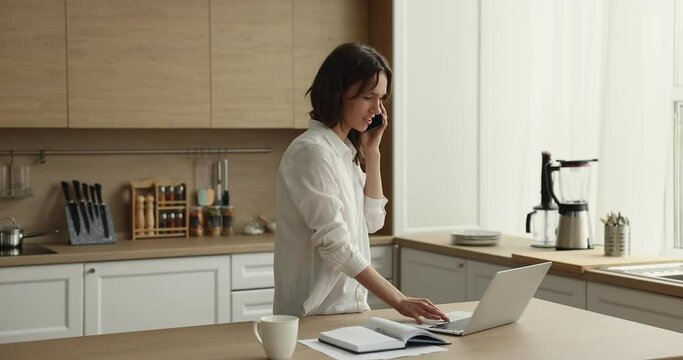 Young attractive woman makes call standing in cozy kitchen using laptop, check e-mail, discuss on-line order. Modern tech, mobile operator usage, distance communication by work, personal talk concept