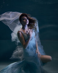 fashionable girl in a beautiful white dress under water in blue light and fluttering fabric in the depths