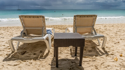 There are two sun loungers and a wicker table on the sandy beach. The boundless turquoise ocean stretches ahead. Foam of waves on the sand. Yachts on the horizon. Tiny silhouettes of swimming people