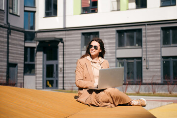 Young woman in coat sits on wooden bench and works with laptop outdoors. Freelance concept.
