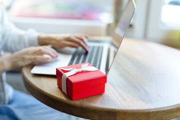 The girl works in a cafe for a laptop and there is a gift nearby. The red gift box lies on the table next to the laptop and the girl.