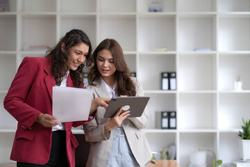 Two beautiful Asian businesswomen standing using digital tablet consulting and analyzing information in office work.