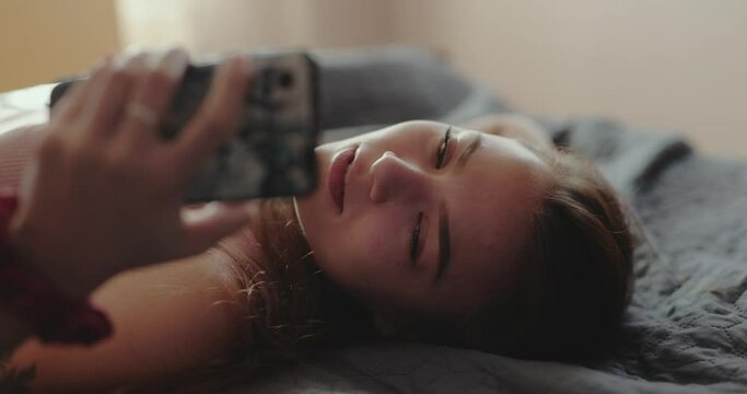 Woman video chatting online talking holding phone lying on a bed at home