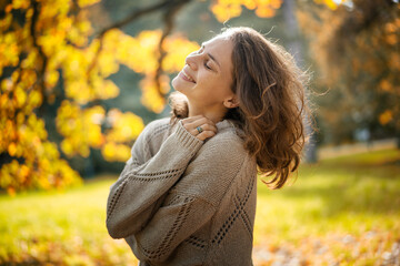 Beautiful smiling caucasian young adult woman in a beige sweater enjoying nature and fresh air walking in the autumn park