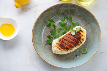 Rustic plate with whole grilled haloumi, fresh parsley and olive oil, high angle view on a light-beige marble background