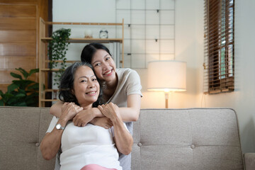 Portrait happy adult daughter and older mother hugging and holding hands, sitting on couch at home,...