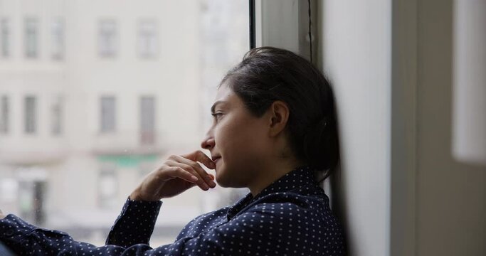 Close up upset young Indian woman sit on windowsill staring out window feels depressed deep in sad thoughts, looking concerned, due to personal troubles, break up, experiences life failure, melancholy
