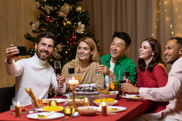 Diverse friends celebrating Christmas and New Year in the evening, sitting at the table, drinking wine and eating, taking a selfie together on the phone near the Christmas tree.