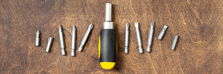 A screwdriver with small bits on a brown wooden table. Banner. Top view