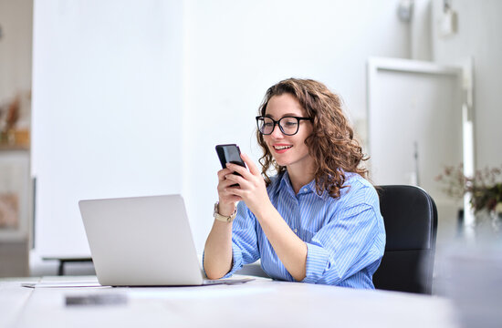 Young Happy Business Woman, Smiling Beautiful Professional Lady Worker Looking At Smartphone Using Cellphone Mobile Cellular Tech Working At Home Or In Office Checking Cell Phone Sitting At Desk.