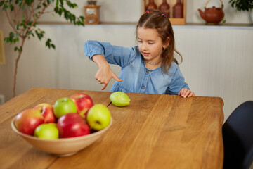 Fototapeta premium Happy little girl twist the pear, the bowl with variety fruits on table at home