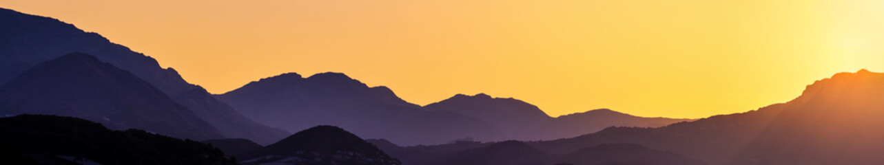 Panoramic view of mountain layers on golden sunset. Silhouettes of hight hills with gradient sunlight. Wide mountain landscape.