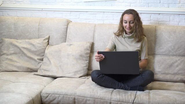 irl blogger communicates with her subscribers. The student calls the teacher. The family is in quarantine. Beautiful cheerful girl with black laptop sitting on sofa in living room at home