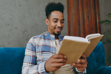 Bottom view young fun man wear casual shirt sits on blue sofa couch read novel stories book stay at home hotel flat rest relax spend free spare time in living room indoors grey wall. People concept.