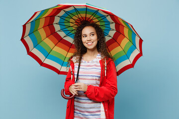 Young minded happy woman of African American ethnicity 20s she wear red jacket hold colorful umbrella look aside isolated on plain pastel light blue cyan background. Wet fall weather season concept.