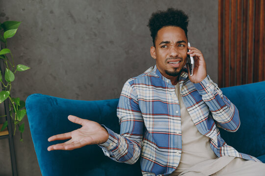 Young Sad Man 20s Wear Casual Shirt Sits On Blue Sofa Couch Talk On Mobile Cell Phone Stay At Home Hotel Flat Rest Relax Spend Free Spare Time In Living Room Indoors Grey Wall. People Lounge Concept.