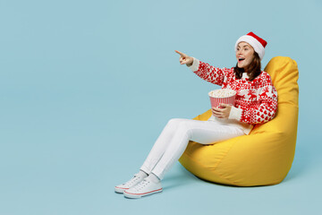 Full body young merry woman wear red knitted sweater Santa hat posing sit in bag chair hold popcorn...