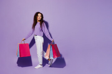 Full body side view young woman wear pullover hold in hand paper package bags after shopping walking go look aside isolated on plain pastel light purple background. Black Friday sale buy day concept.