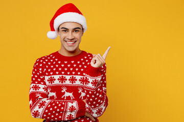 Merry young man in knitted christmas sweater Santa hat posing point index finger aside indicate on...