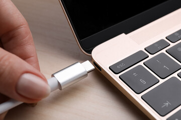 Woman plugging USB cable with type C connector into laptop port on light wooden table, closeup
