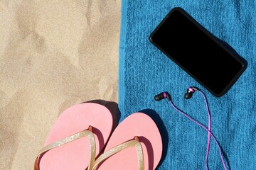 Soft blue beach towel with flip flops, smartphone and earphones on sand, flat lay. Space for text