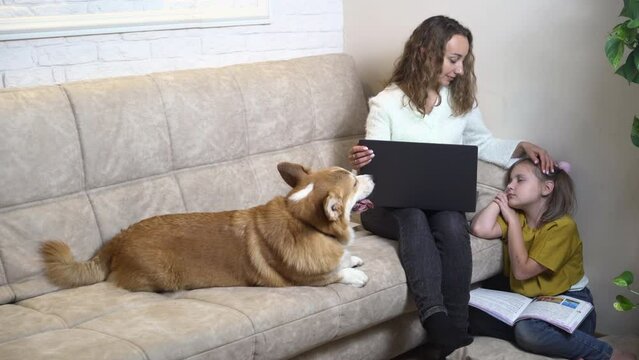 A Young Woman In A White Cardigan Sits At Home On The Sofa And Looks At The Laptop Screen. The Girl Is Shopping Online. A Girl Of 10 Years Old Is Reading A Book, 