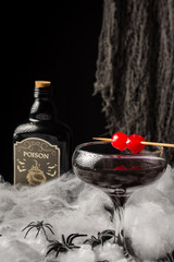 Close-up of glass of Halloween black widow cocktail with red cherries, spiders and dark bottle on spider web, black background, vertical