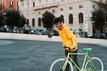 Happy young African man having fun with bike in the city while listening music with headphones -...