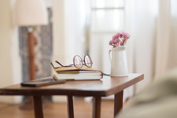 Bedside table with eyeglasses, books, flowers and phone on it. Medium shot of beautiful brown bedside cabinet located in cozy bedroom. Interior, furniture concept