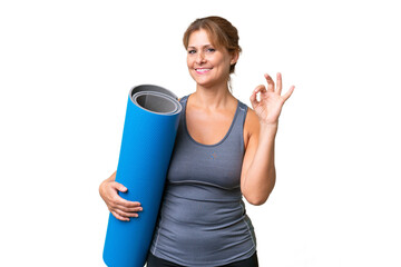Middle-aged sport woman going to yoga classes while holding a mat over isolated background showing ok sign with fingers © luismolinero