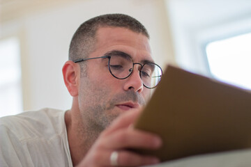 Cheerful man lying on bed in bedroom and reading book. Close-up shot of focused man in eyeglasses...