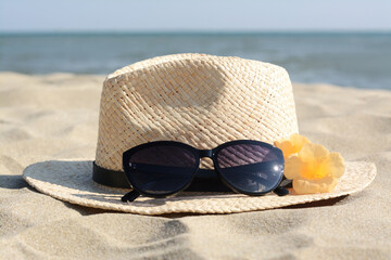 Hat with beautiful sunglasses and flowers on sand near sea, closeup