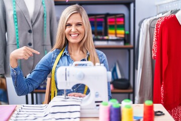 Naklejka na ściany i meble Blonde woman dressmaker designer using sew machine looking confident with smile on face, pointing oneself with fingers proud and happy.