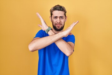Hispanic man with beard standing over yellow background rejection expression crossing arms doing...