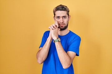 Hispanic man with beard standing over yellow background holding symbolic gun with hand gesture,...