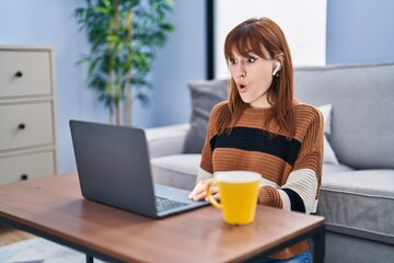 Young beautiful woman using computer laptop doing video call scared and amazed with open mouth for surprise, disbelief face