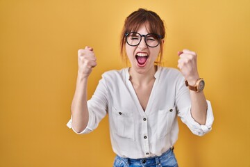Young beautiful woman wearing casual shirt over yellow background angry and mad raising fists frustrated and furious while shouting with anger. rage and aggressive concept.