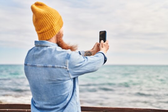 Young redhead man standing on back view make photo by smartphone at seaside