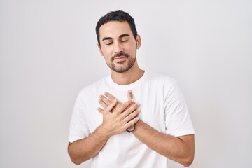 Handsome hispanic man standing over white background smiling with hands on chest with closed eyes and grateful gesture on face. health concept.