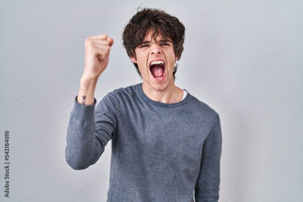 Poster young man standing over isolated background angry and mad raising fist frustrated and furious while 